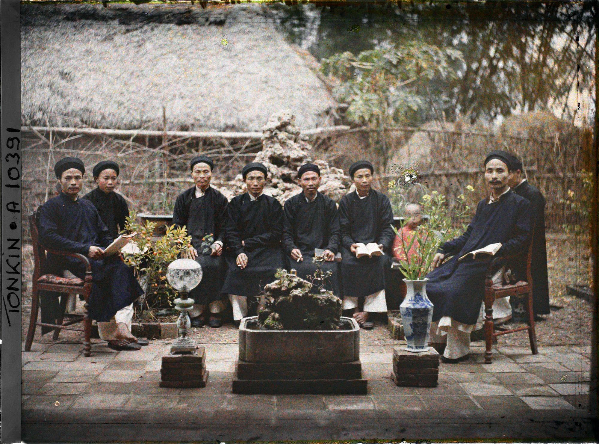 Một nhóm hương chức ngồi trong một sân nhà ở miền Bắc Việt Nam. Sân vườn bài trí chậu quất, bình hoa, gợi không khí về phong quang mời khách ngâm thơ trong vườn nhà trong thơ của Trần Phi Khanh
